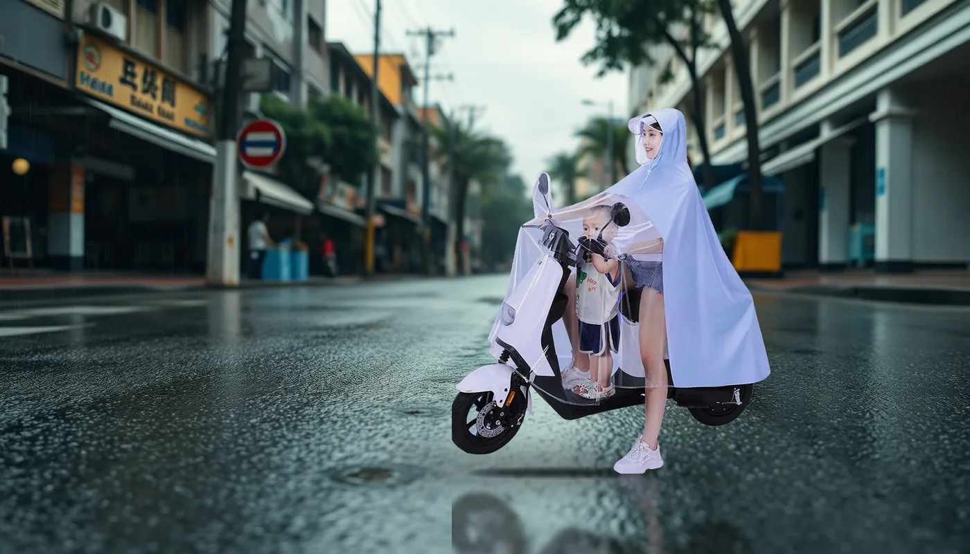 An adult rider and a child on an e-bike on a wet street in the rain, both under one family rain poncho with a clear face visor and a wide front shield.